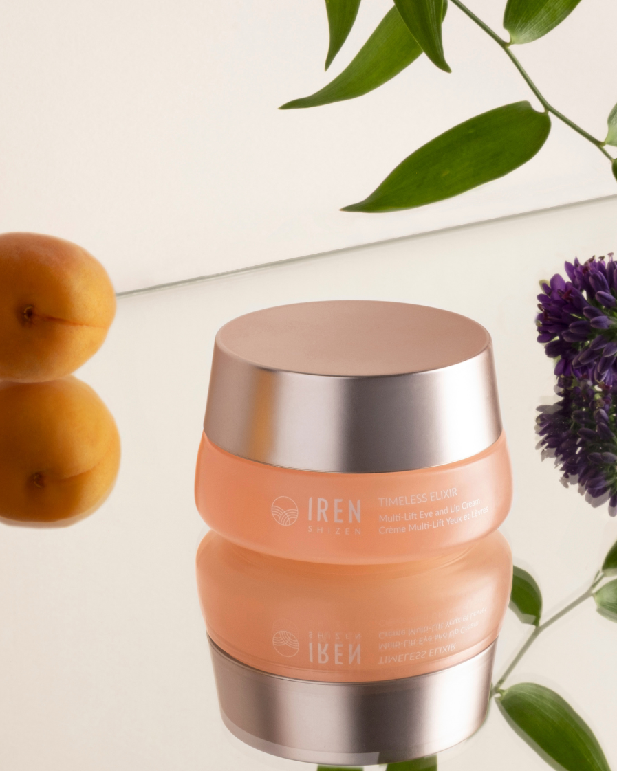 A jar of IREN Shizen TIMELESS ELIXIR Hydrating Eye Skincare Kit, celebrated for its anti-aging benefits, is placed on a reflective surface, surrounded by apricots and green leaves, with a purple flower nearby.