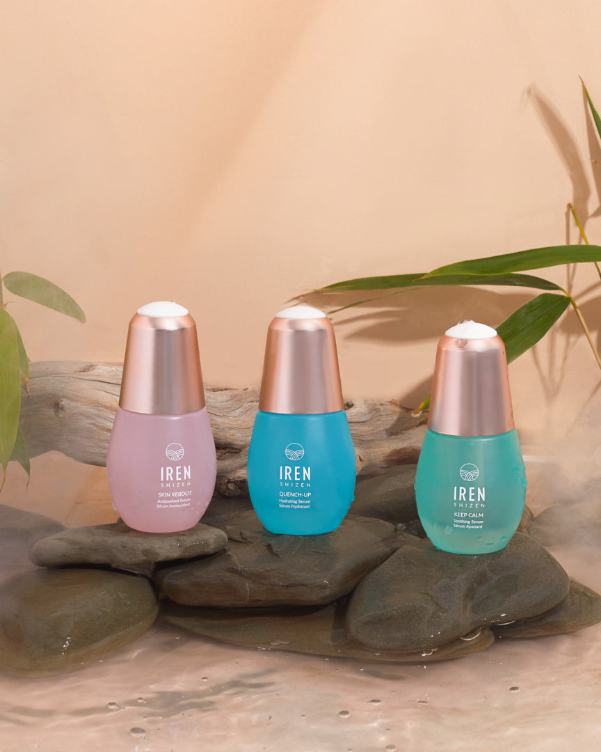 The IREN Shizen 3-SERUM ROUTINE Custom Set, in pastel pink, blue, and turquoise bottles, rests on smooth rocks with bamboo stems and driftwood in the background.