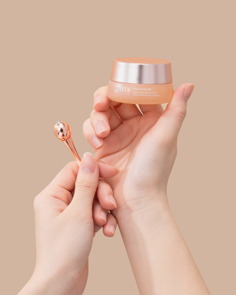 Hands holding a small jar of the TIMELESS ELIXIR Multi-Lift Eye and Lip Cream by IREN Shizen, infused with Bakuchiol and accompanied by a metallic applicator, set against a beige background.