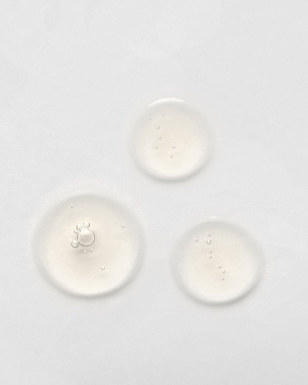 Three small, clear liquid droplets on a plain white surface, possibly from the LIFT & FIRM Anti-Aging Duo Serum Set by IREN Shizen, which contains Argireline Peptide.