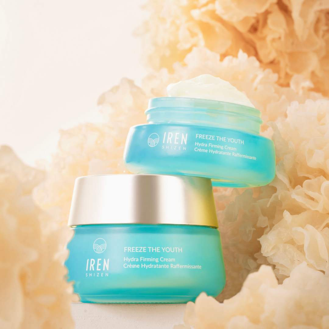 Image of two blue jars of FREEZE THE YOUTH Hydra Firming Cream by IREN Shizen with the age-defying formula, placed among light brown decorative fibers. The cream, enriched with snow mushroom extract, is visible in the top jar, which is open, while the bottom jar is closed.